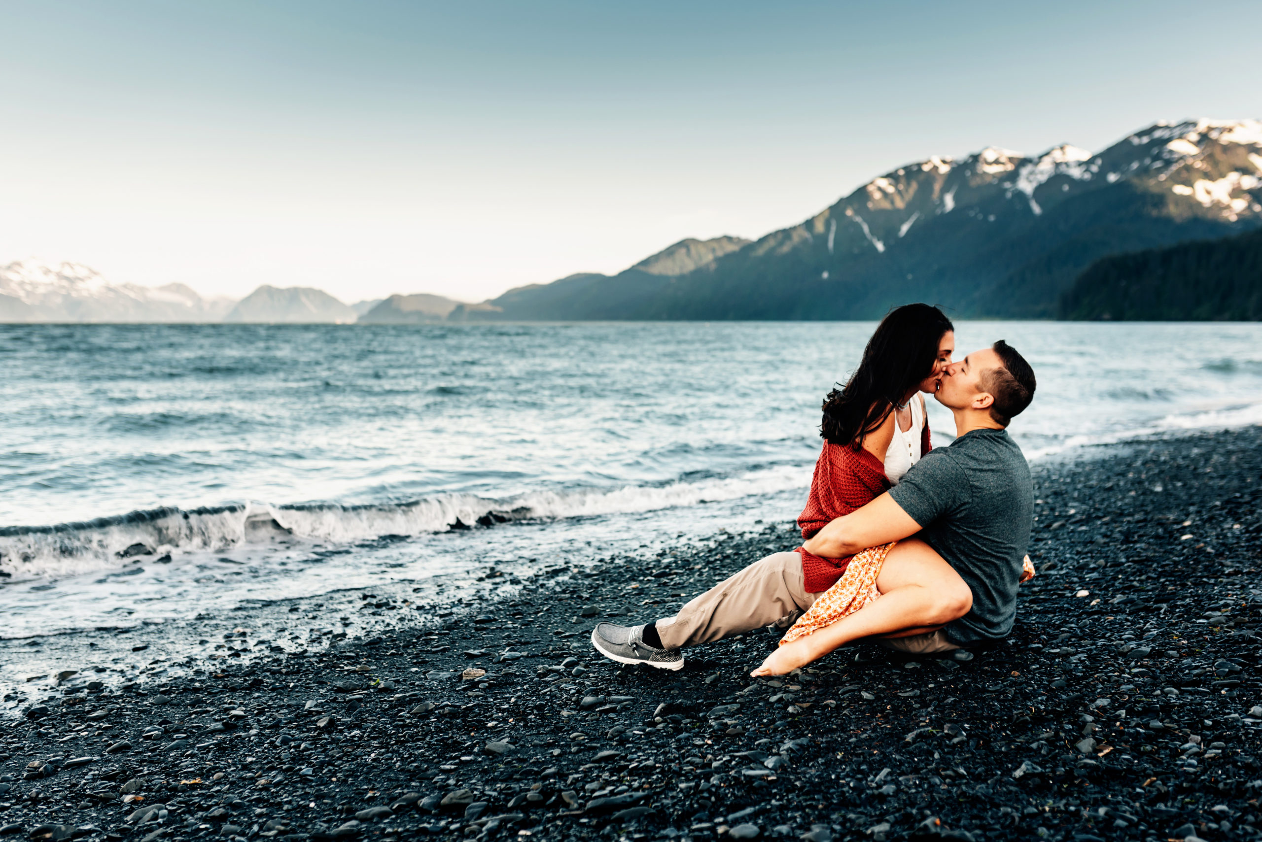 Woman straddling husband. Kissing one another while sitting on a black rock beach along the coast of Alaska.